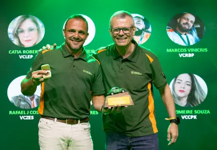 Dois homens de camisa verde sorrindo com o troféu, diante do telão com os rostos dos premiados