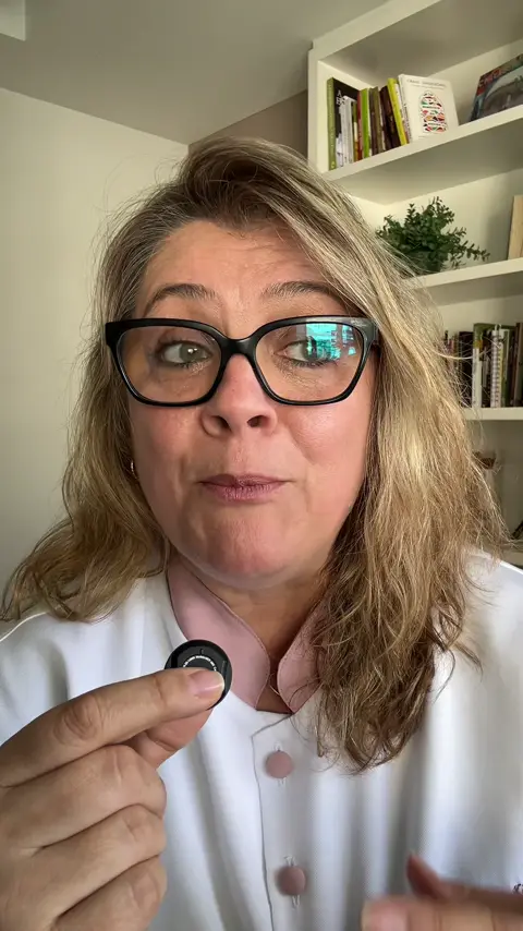 Dra. Ana Luiza, de jaleco branco, fala para a câmera segurando o microfone de lapela